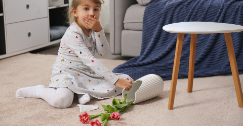 Een klein meisje schrikt en heeft haar hand voor haar mond omdat de vaas met bloemen gevallen en gebroken is